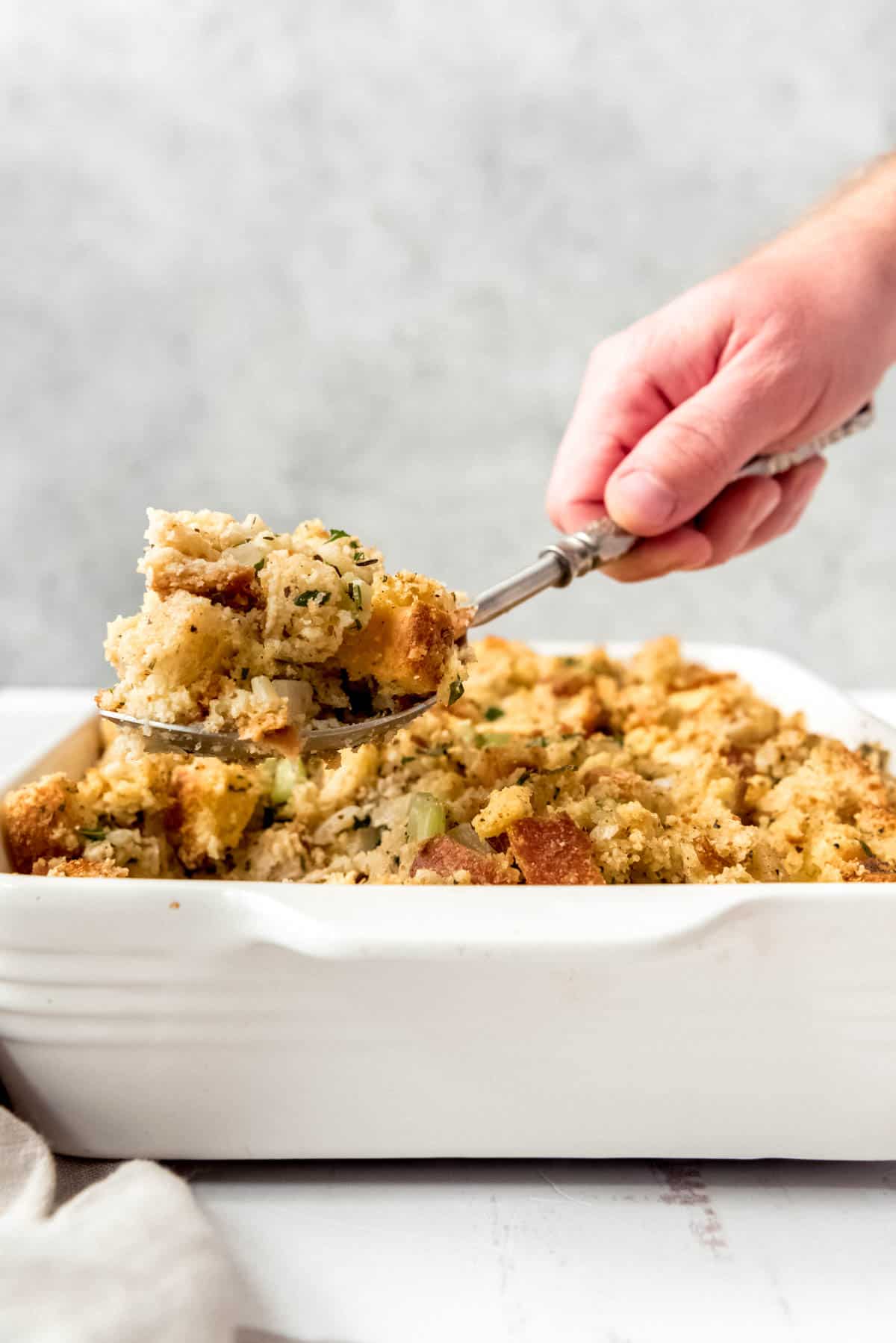 An image of a hand lifting a scoop of cornbread dressing out of a white baking dish.