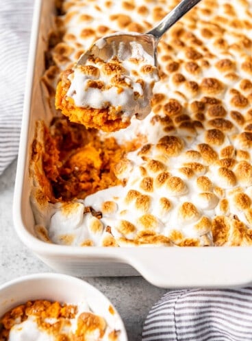 Sweet potato casserole with marshmallows in a white baking dish with a spoon scooping some out of the pan.