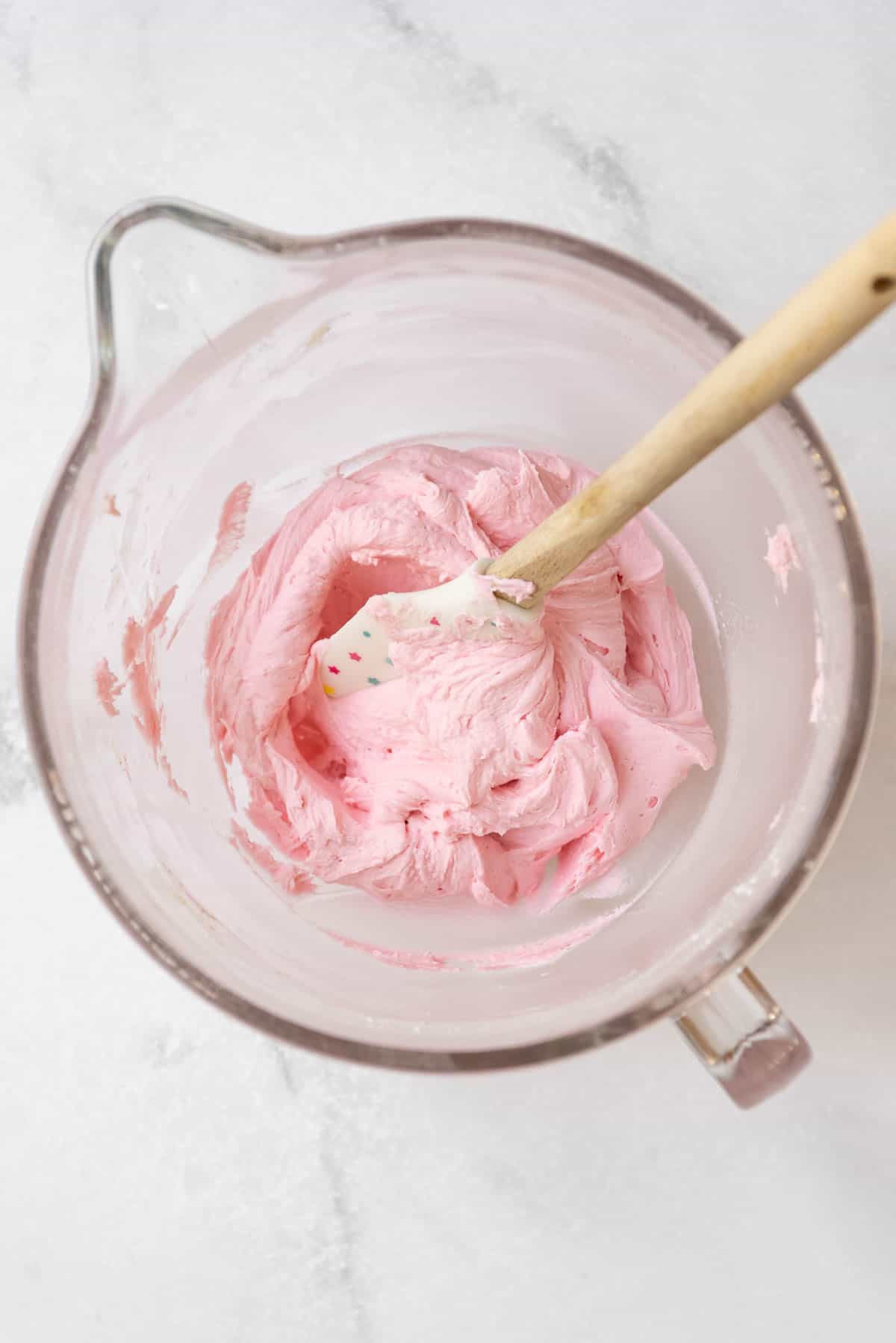 A spatula in a lass glass mixing bowl filled with pink frosting.