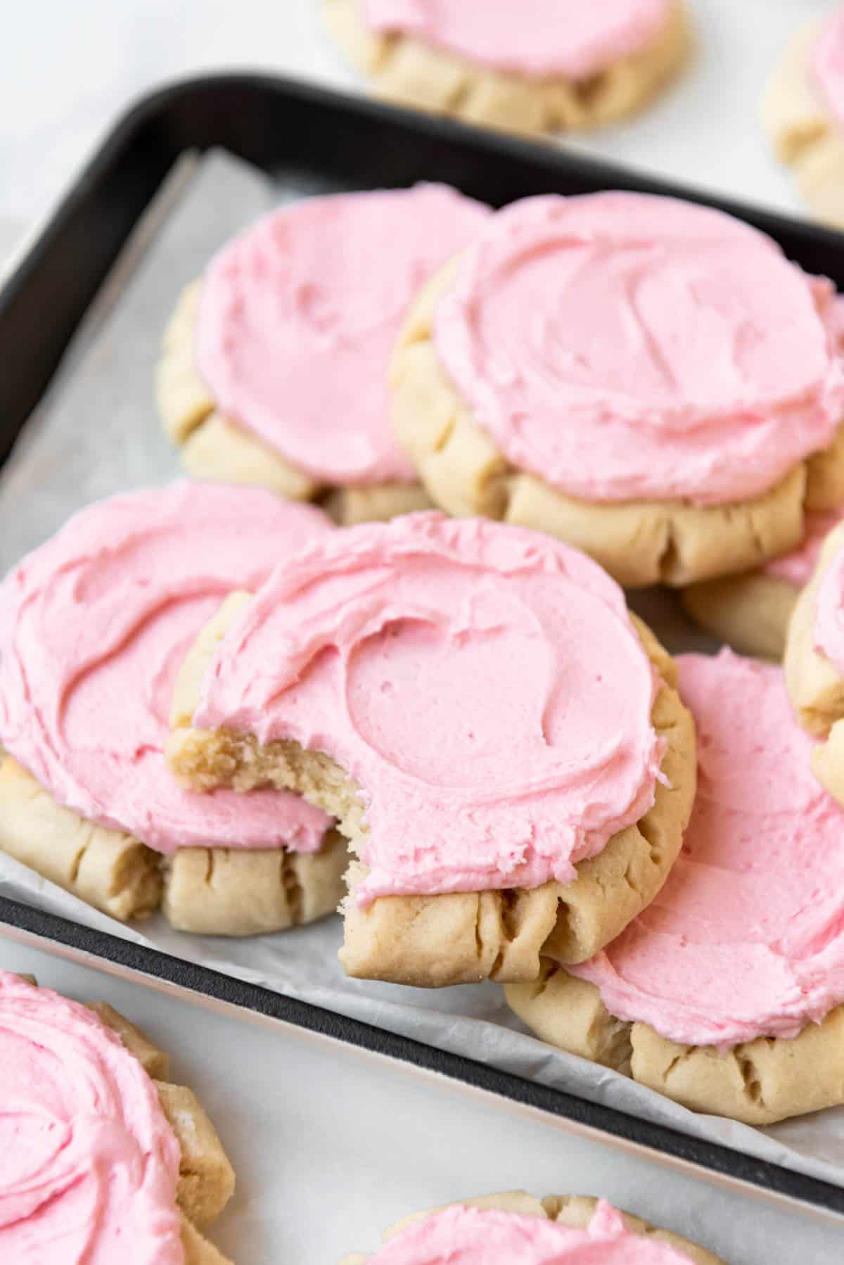 A pink frosted sugar cookie with a bite taken out of it with more sugar cookies on an angled serving plate.