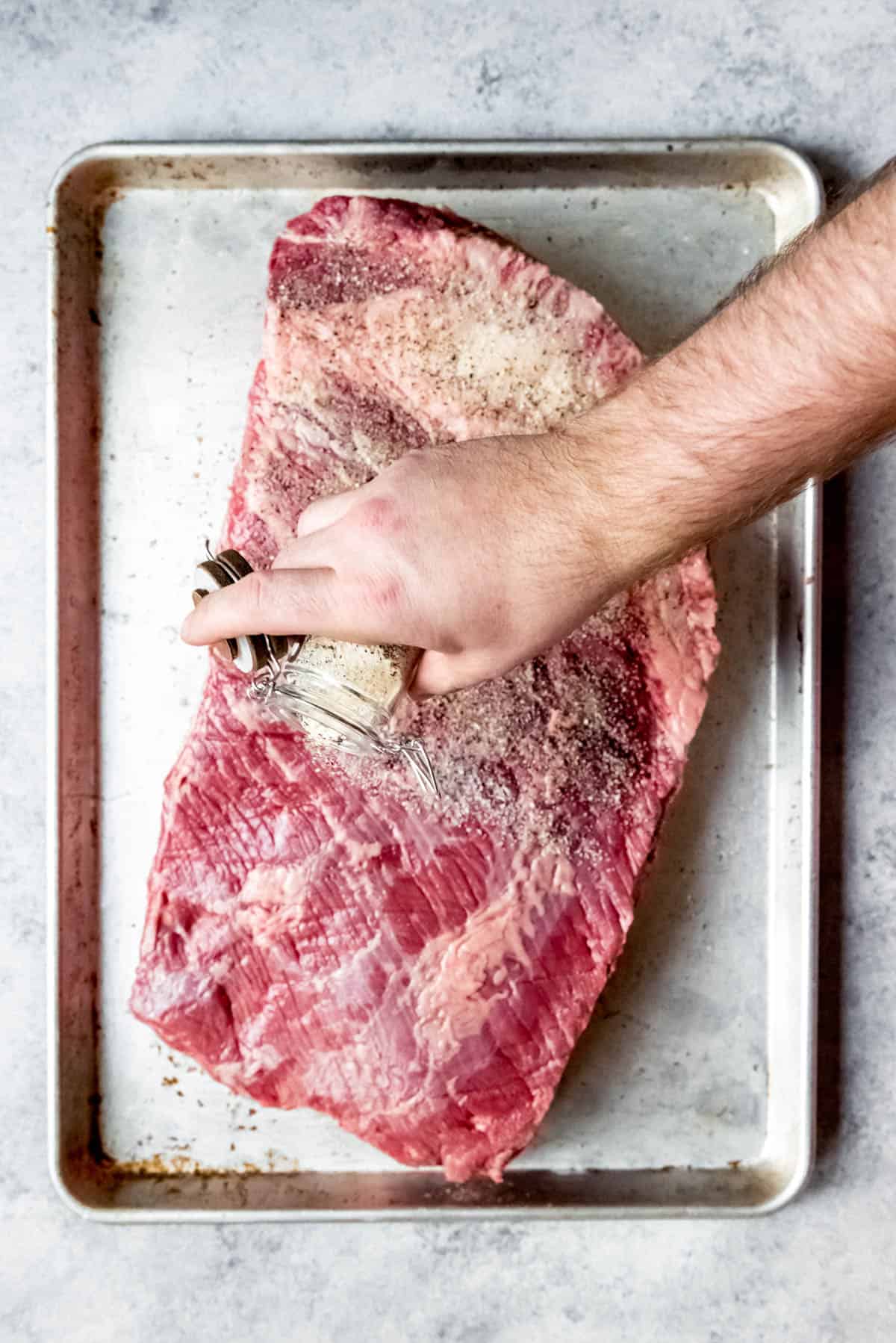 An image of a person's hand sprinkling a salt, pepper, and garlic rub over a trimmed beef brisket.