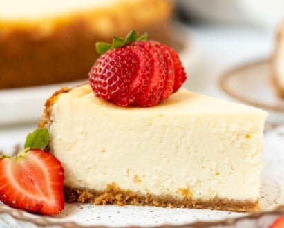 A slice of the best cheesecake on a plate with strawberries in front of the rest of the cheesecake.