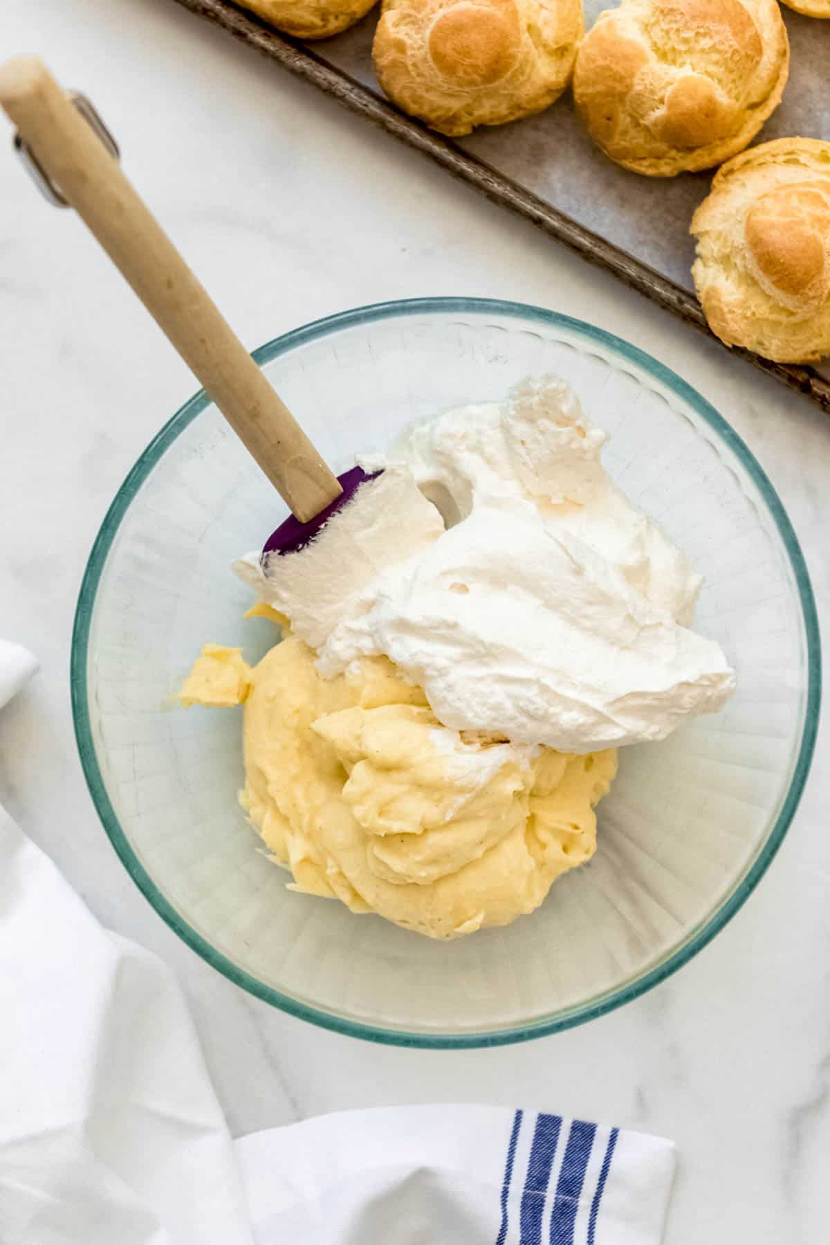 A glass bowl with pastry cream and whipped cream next to cream puff shells.