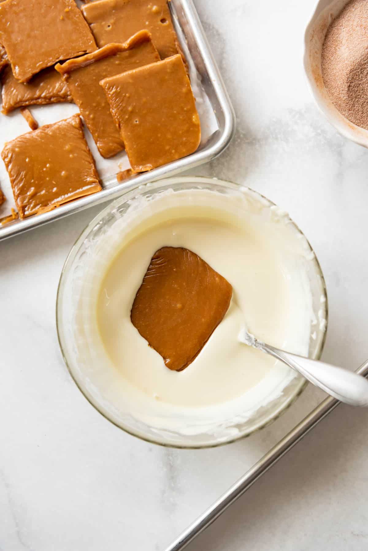 An image of a square piece of toffee getting dipped into a bowl of white chocolate.