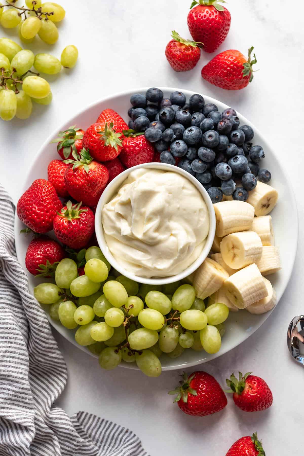 A bowl of fruit dip on a plate with strawberries, grapes, bananas, and blueberries.