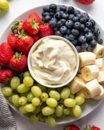 A bowl of fruit dip on a plate with strawberries, grapes, bananas, and blueberries.
