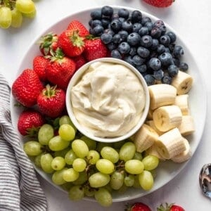 A bowl of fruit dip on a plate with strawberries, grapes, bananas, and blueberries.