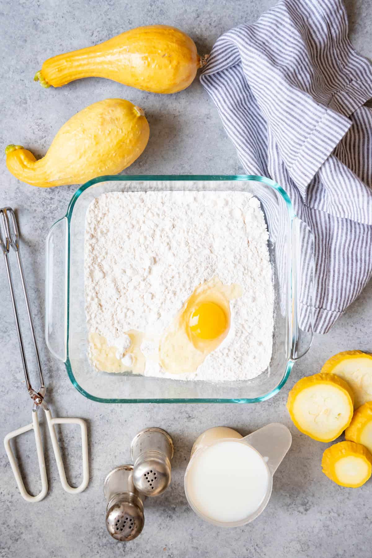 An image of egg in flour mixture in baking dish sourrounded by other ingredients and tools needed.