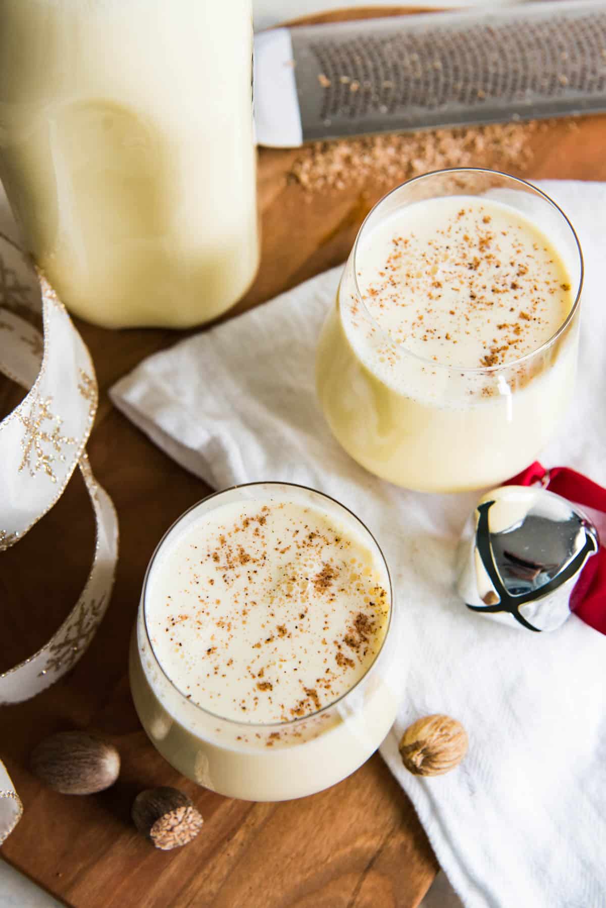 Top view of two glasses of nutmeg topped with grated nutmeg with a bell  on a white towel, a gold ribbon and a bottle of eggnog also in the frame.