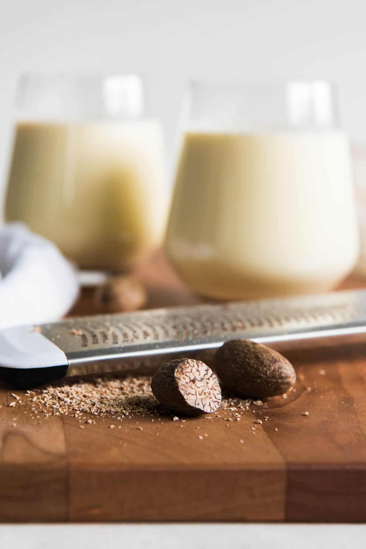 Close up of a zester, some nuts, and two glasses of eggnog in the background on a chopping board. 