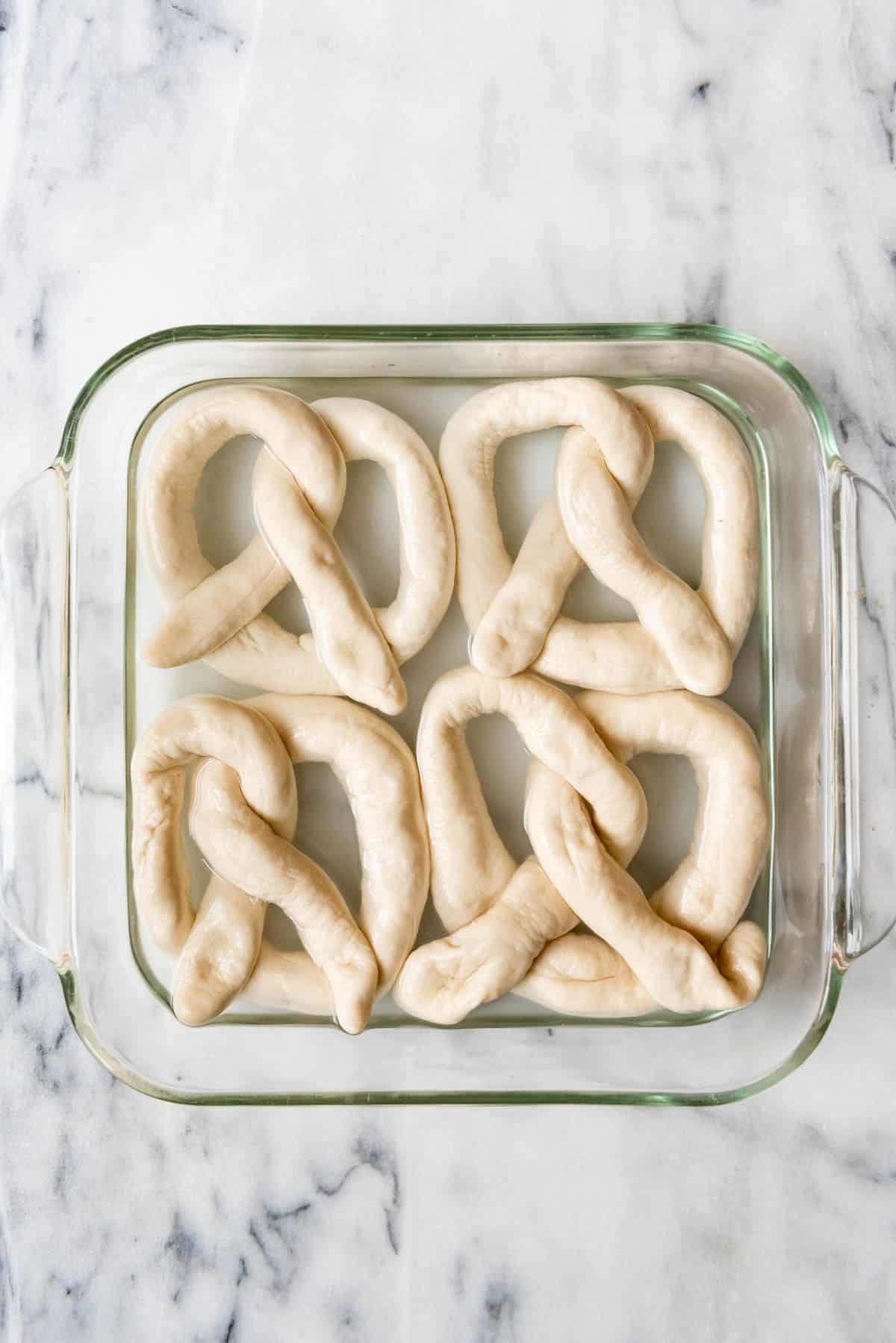 An image of 4 pretzels soaking in a baking soda bath in square dish.