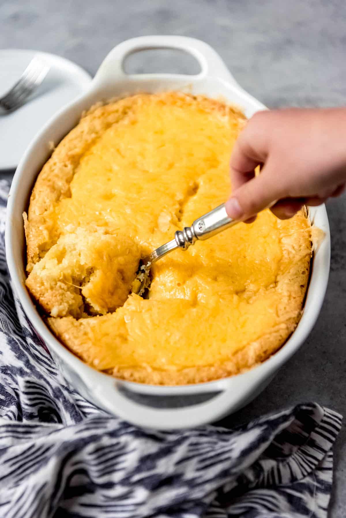 An image of a hand taking a big scoop of corn pudding out of a casserole dish.