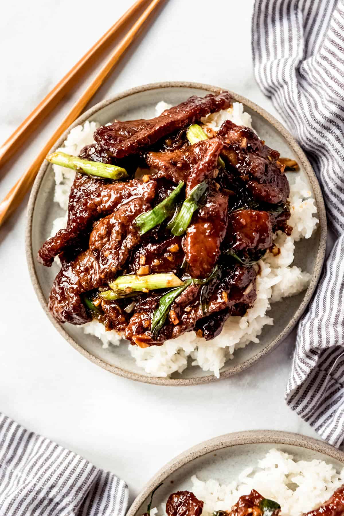 Mongolian Beef in bowl on top of rice with chopsticks and a striped towel on the sides of the bowl.