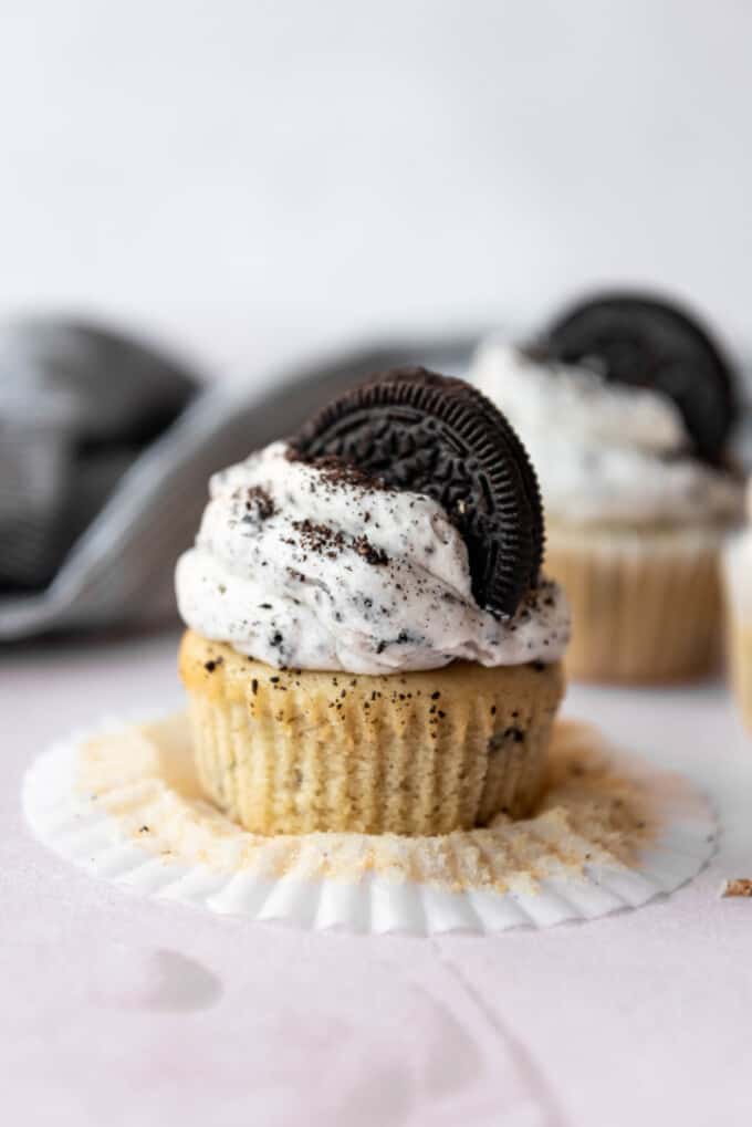 An Oreo cookie cupcake with cookies and cream frosting and an Oreo on top, sitting on worktop with the cupcake case peeled off.