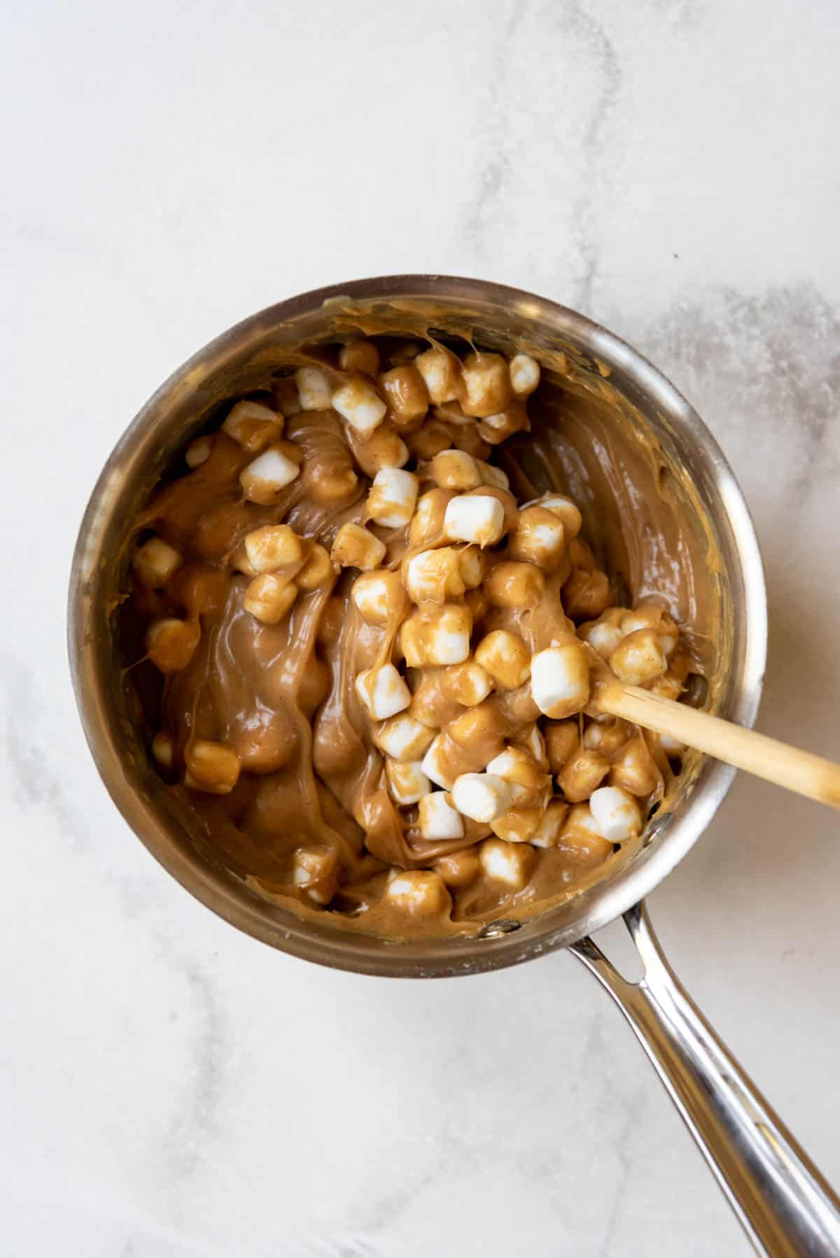 A wooden spoon being used to stir marshmallows into a melted peanut butter mixture in a medium pan.