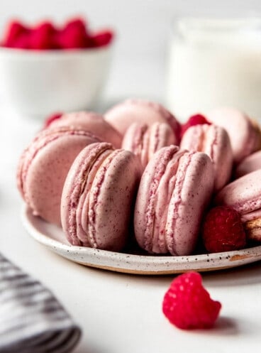 Fresh red raspberries around a plate of nicely arranged pink Raspberry macarons.
