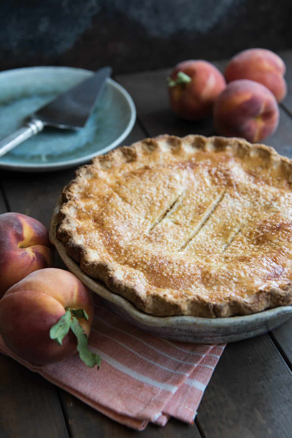 A Southern Peach Pie in a pie plate next to fresh whole peaches.