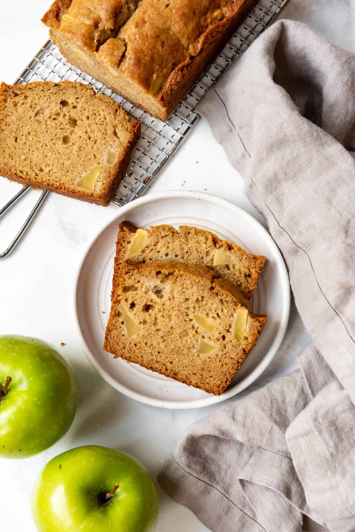 Sliced apple ginger quick bread on a plate next to the rest of the loaf and two green apples.
