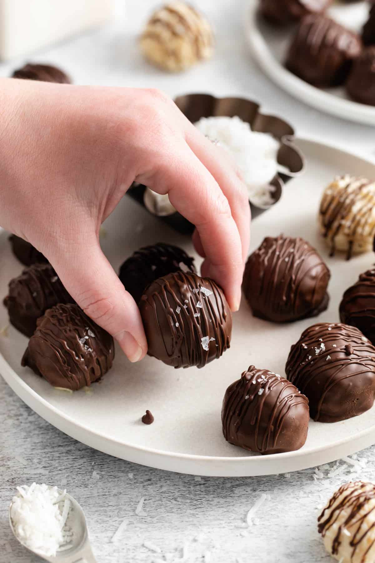 A hand picking up a chocolate coconut ball from a plate.