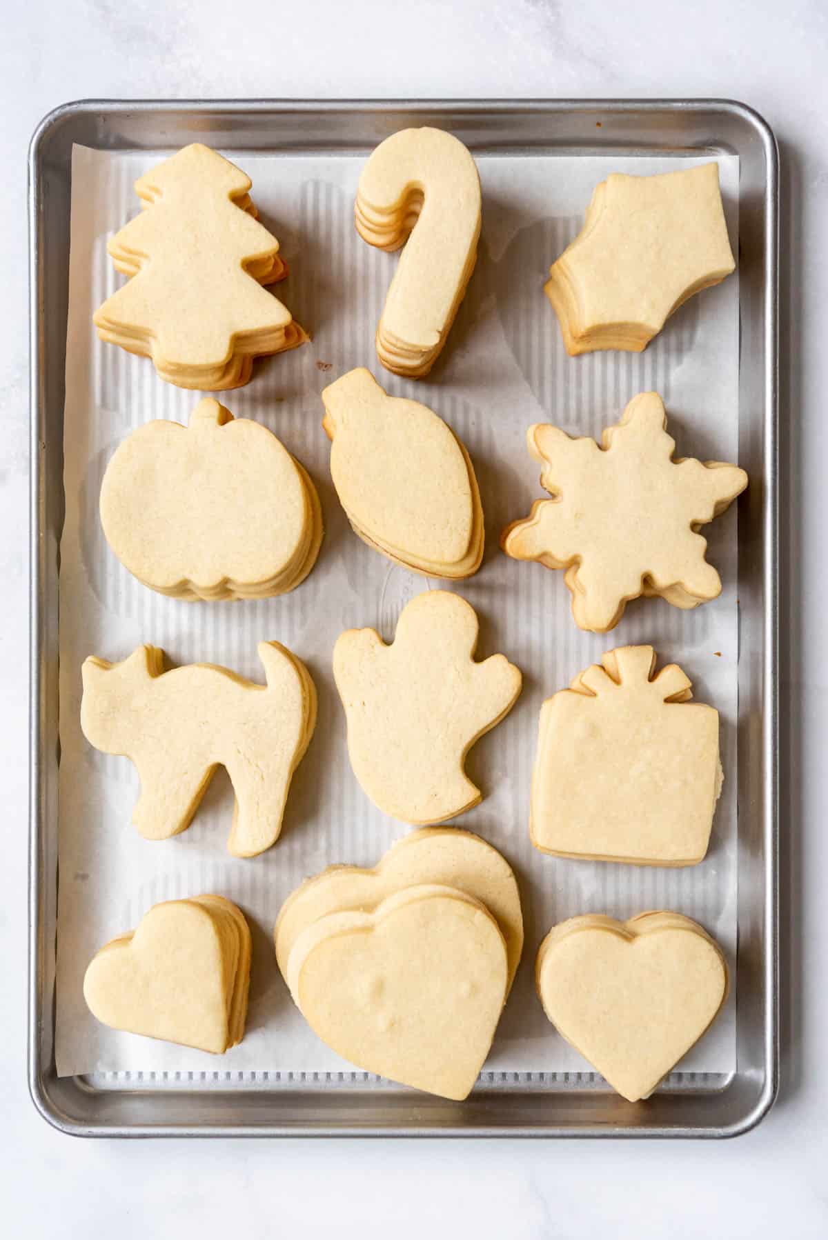 An overhead image of a baking sheet filled with stacks of unfrosted sugar cookies cut out in different holiday shapes.