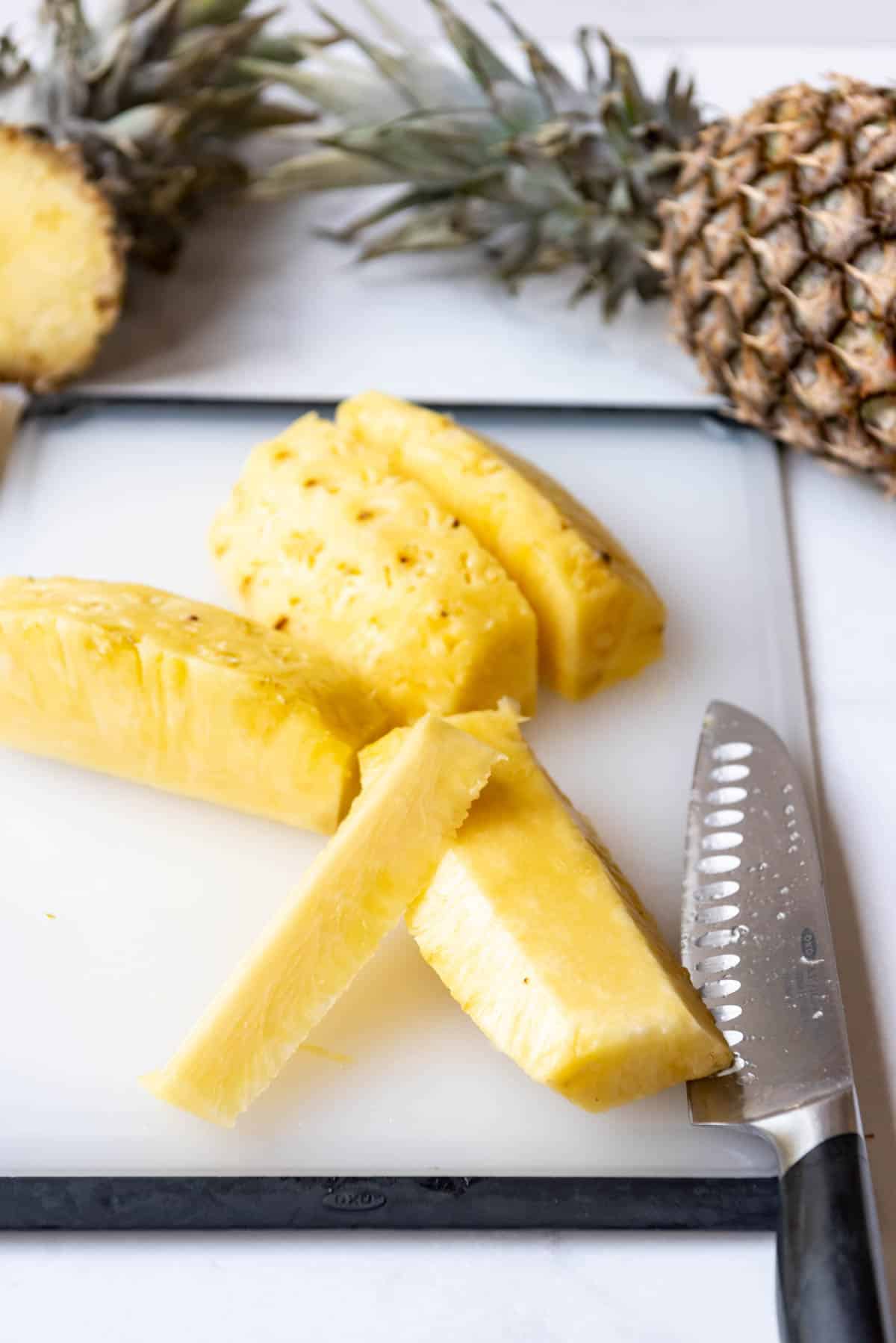 A knife on a cutting board with pineapple cut into wedges in front of a whole pineapple.