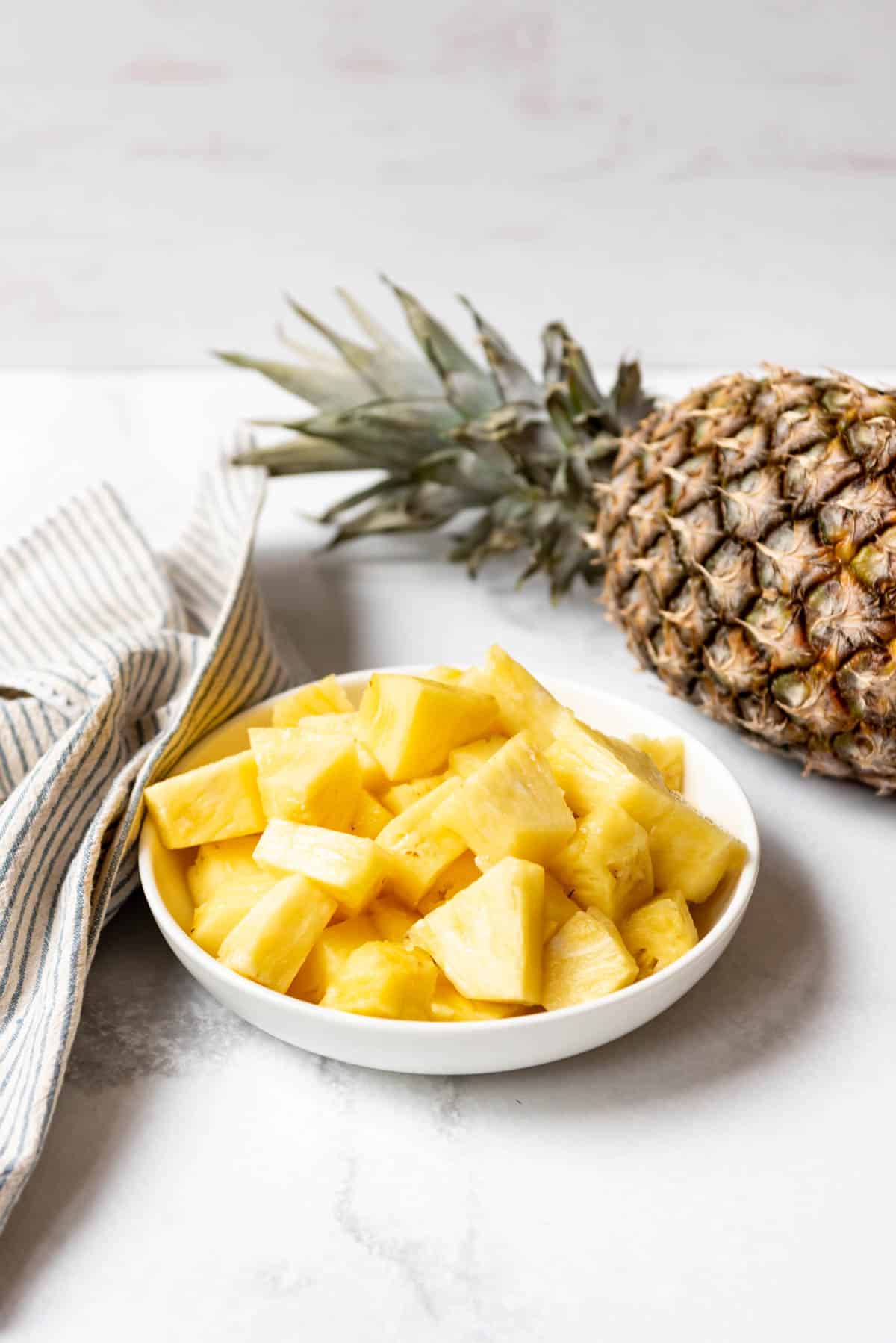 A bowl of fresh pineapple chunks in front of a whole pineapple.