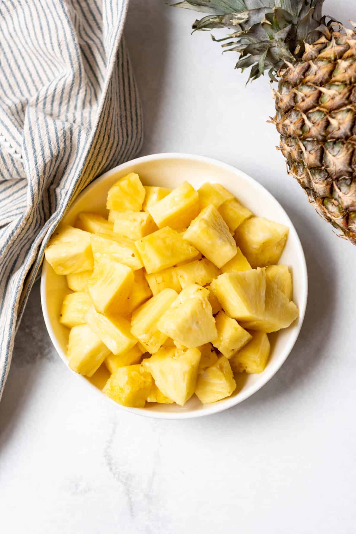 An overhead image of fresh pineapple chunks next to a whole pineapple and striped cloth.