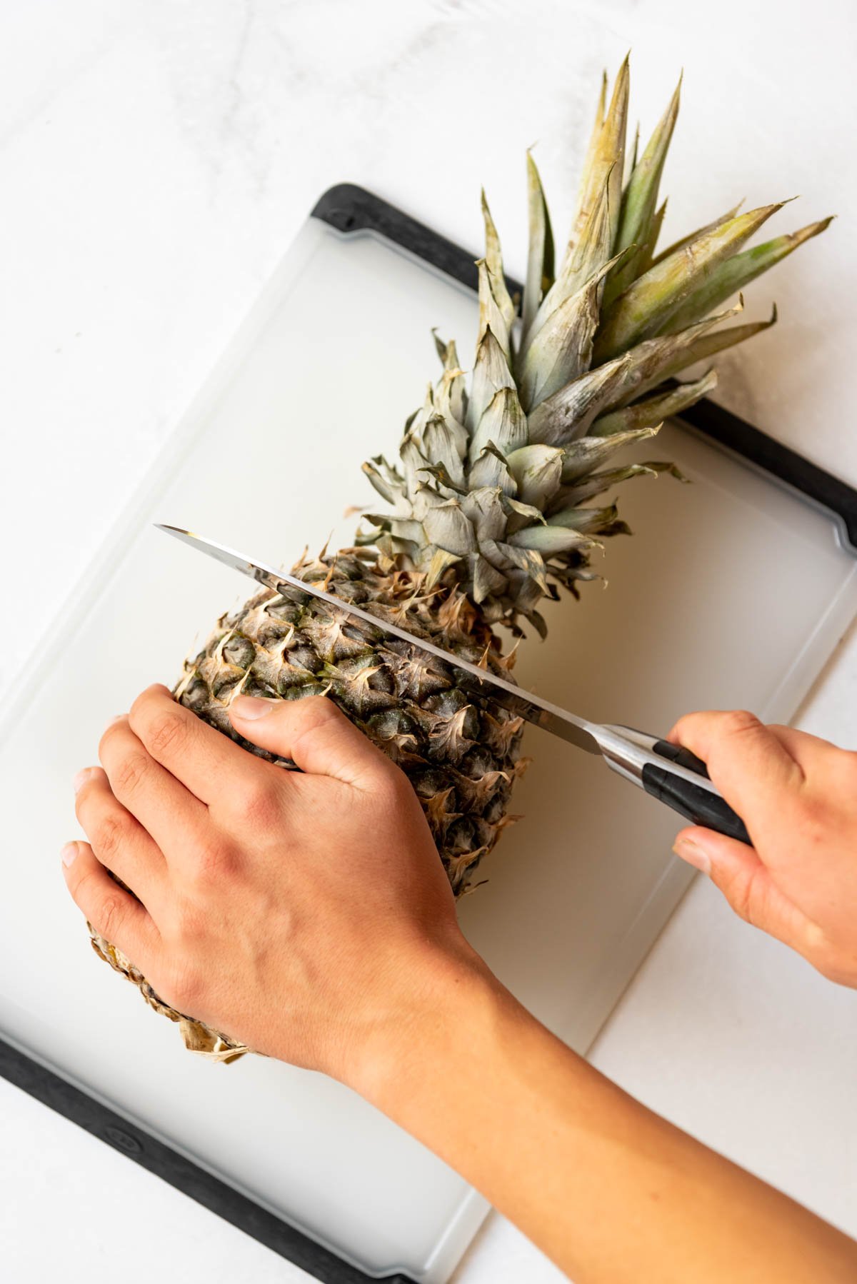 A hand using a knife to cut off the top or crown of a fresh pineapple.