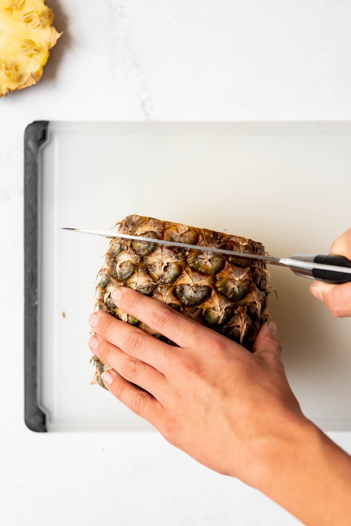 Hands holding a knife to slice a fresh pineapple on a cutting board.