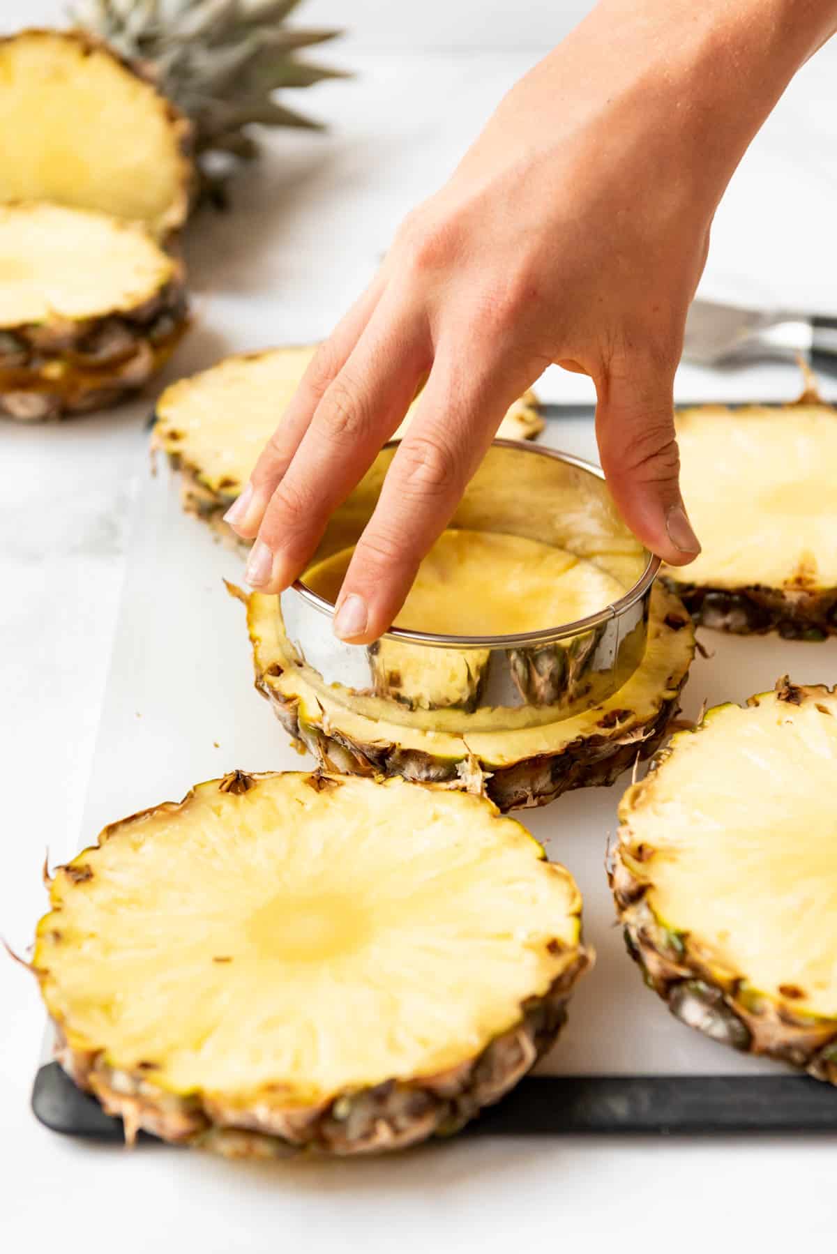 A hand pressing a large circular metal cutter to cut out a pineapple ring from a slice of fresh pineapple.