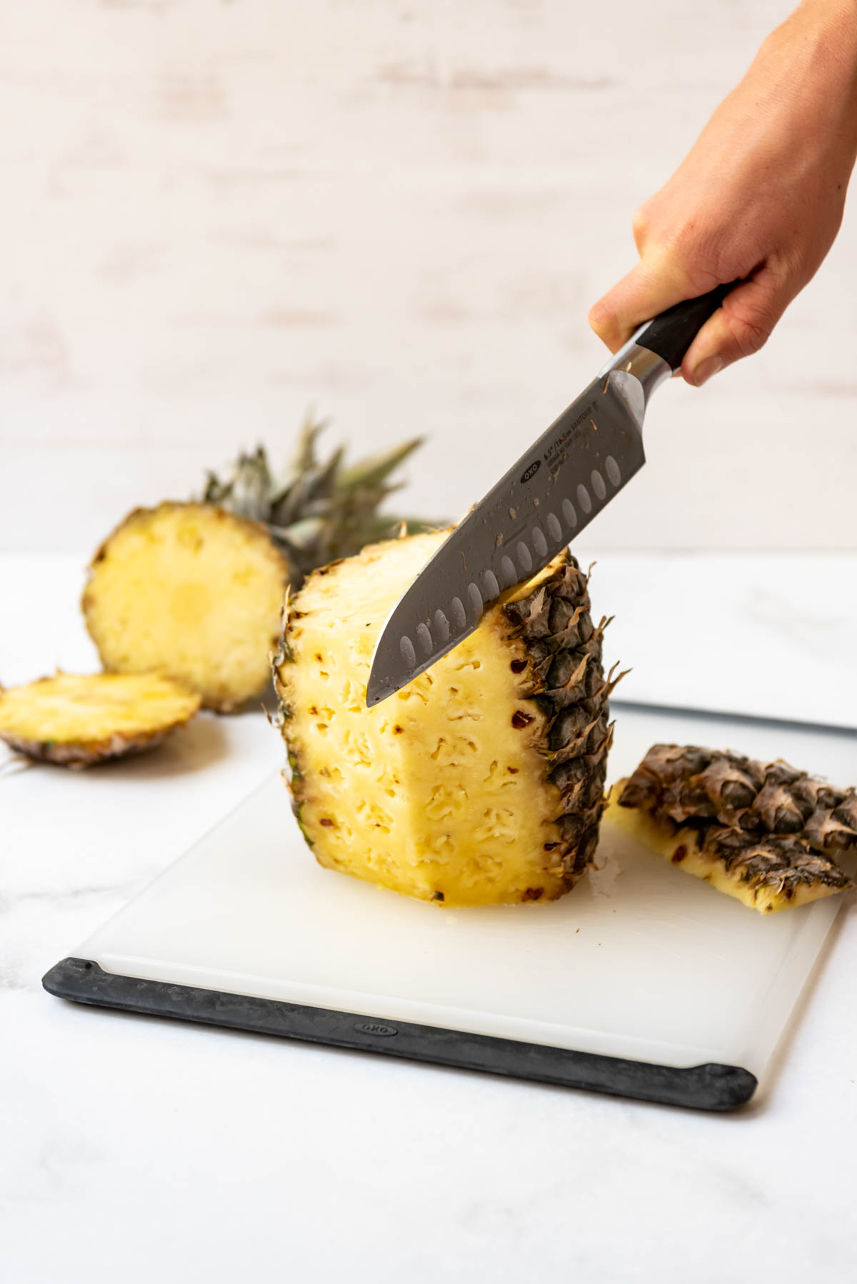 A knife being used to remove the tough outer skin of a fresh pineapple on a cutting board.