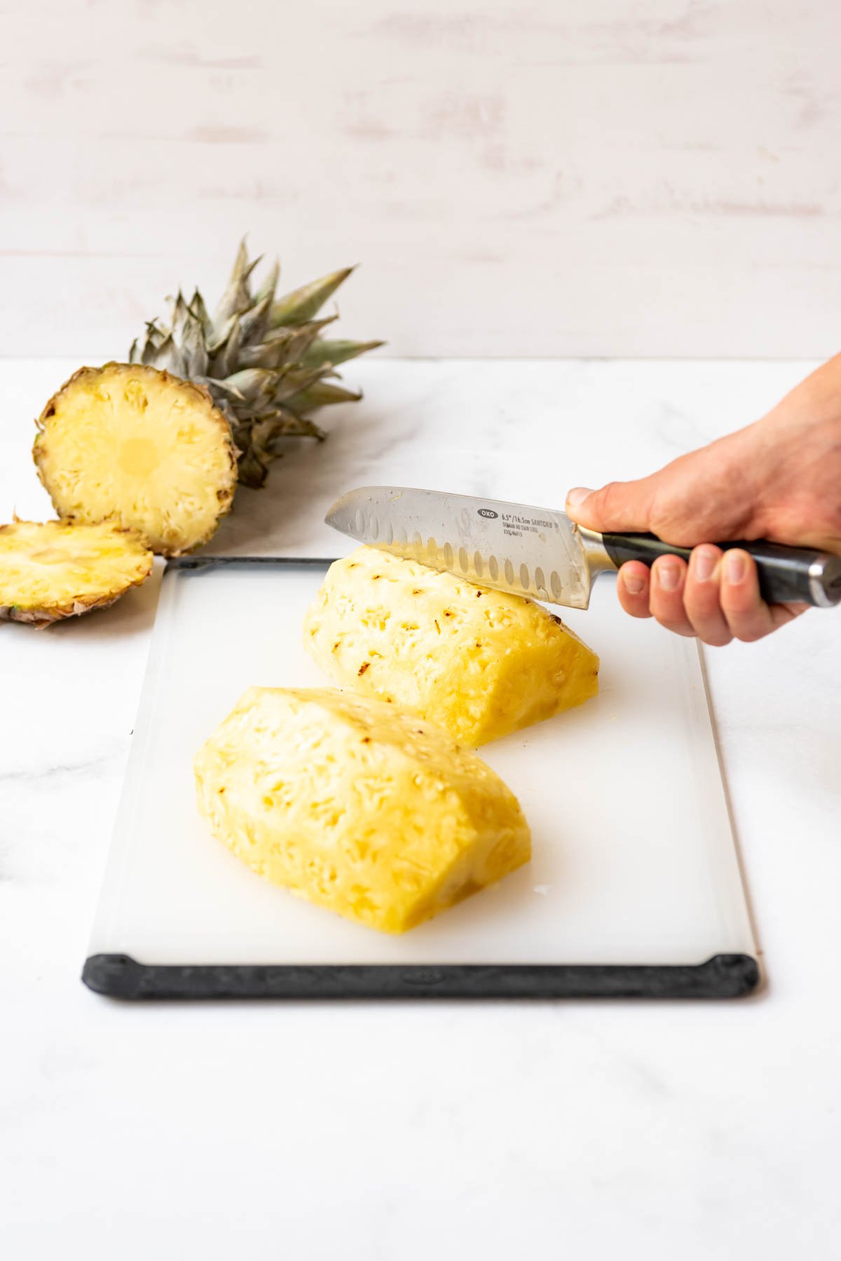 A hand holding a knife to cut pineapple on a cutting board.
