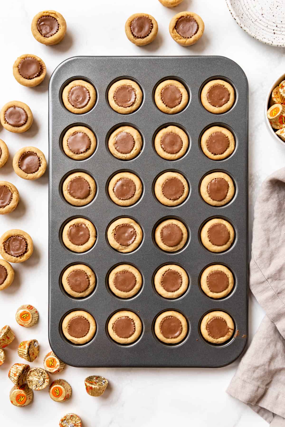 An overhead image of peanut butter cup cookies in a mini muffin pan with more cookies and miniature peanut butter cups scattered around it.