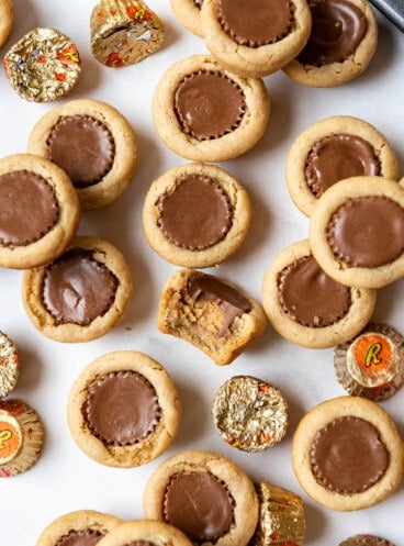Peanut Butter Cup Cookies with a bite taken out of one of them and some wrapped mini Reese's peanut butter cups scattered around them.