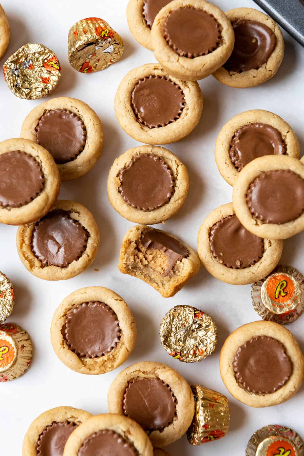 Peanut Butter Cup Cookies with a bite taken out of one of them and some wrapped mini Reese's peanut butter cups scattered around them.