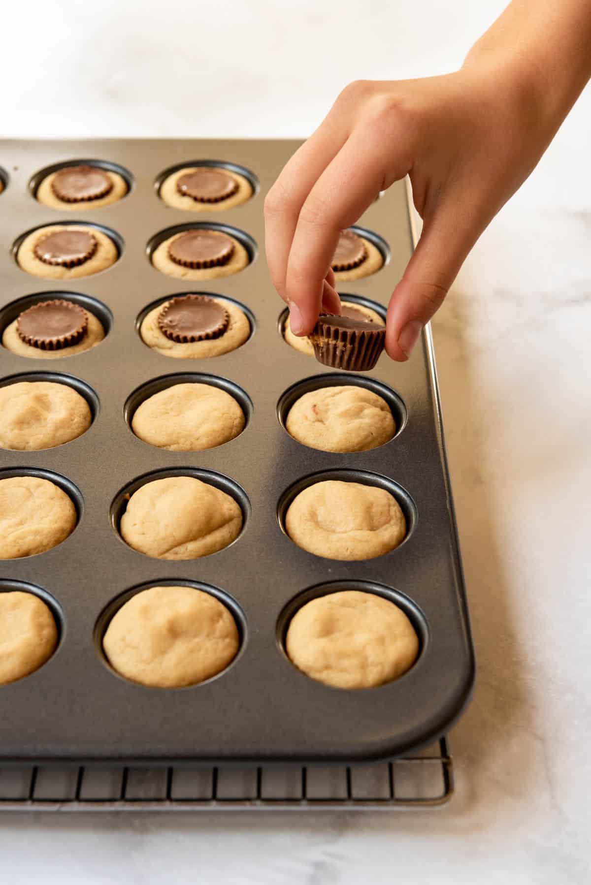 A hand holding an unwrapped mini Reese's peanut butter cup over a tray of baked peanut butter cookies.