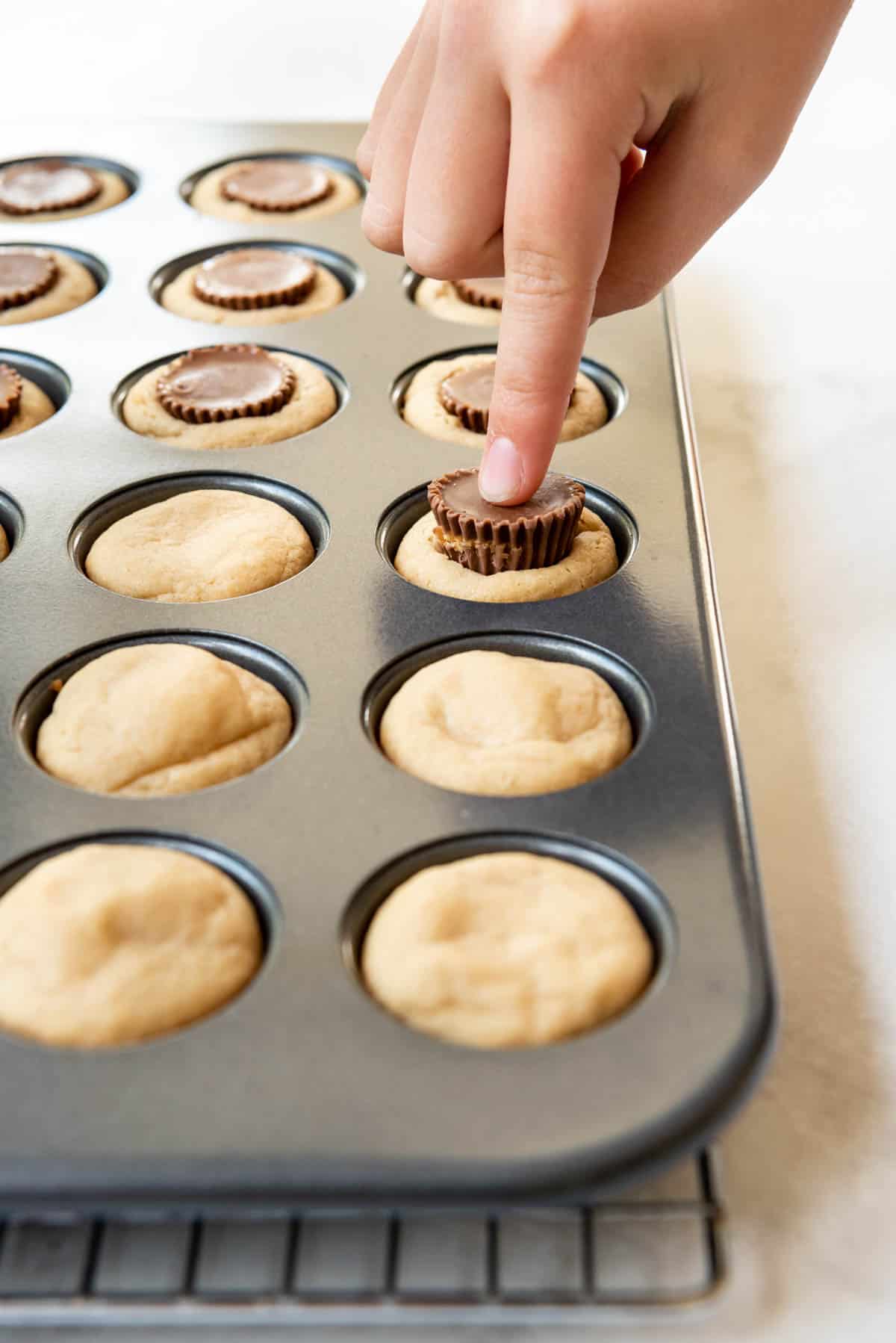 A finger pushing an unwrapped mini Reese's peanut butter cup into baked peanut butter cookie dough in a mini  muffin pan.