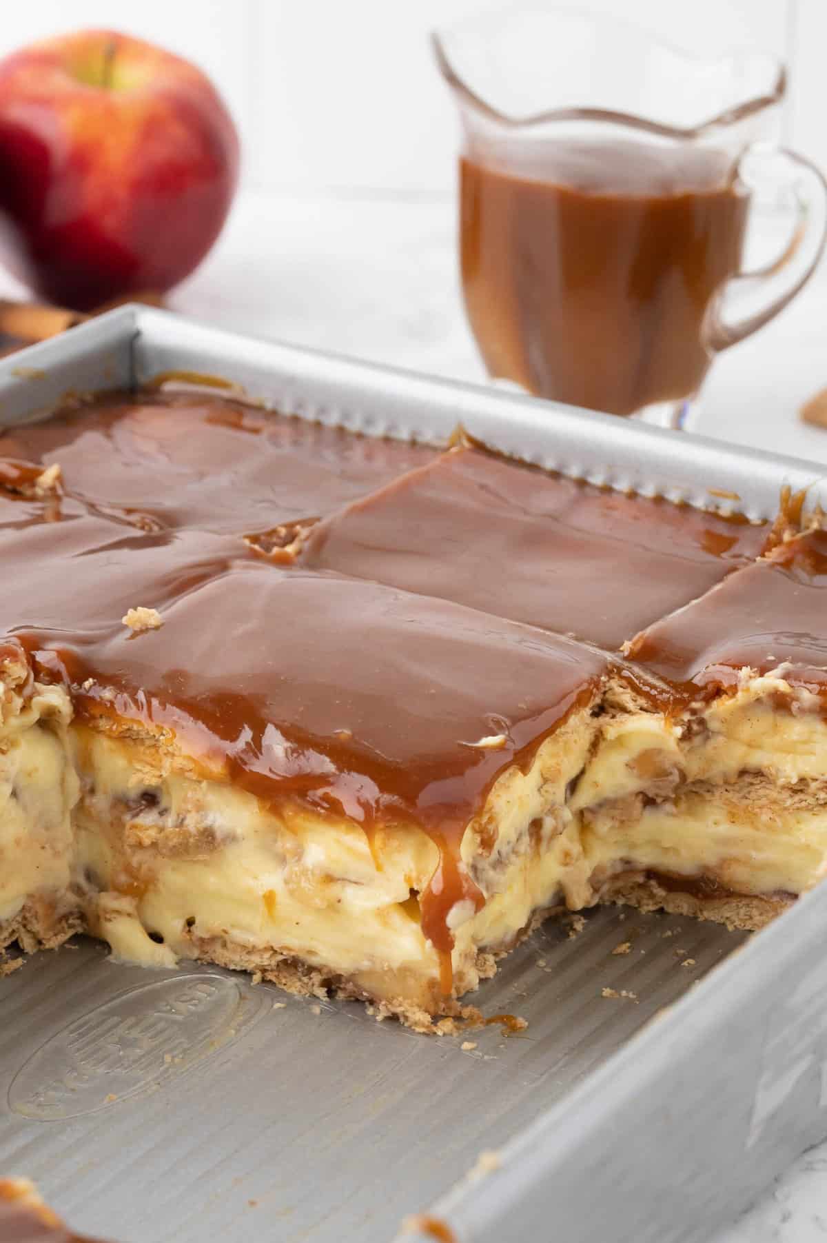 Slices of caramel apple eclair cake in a baking dish in front of a glass jar of caramel sauce and an apple.