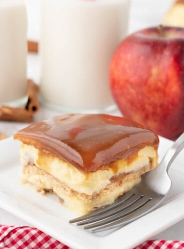 A slice of caramel apple eclair cake on a white plate with a red gingham napkin and a glass of milk next to an apple behind it.