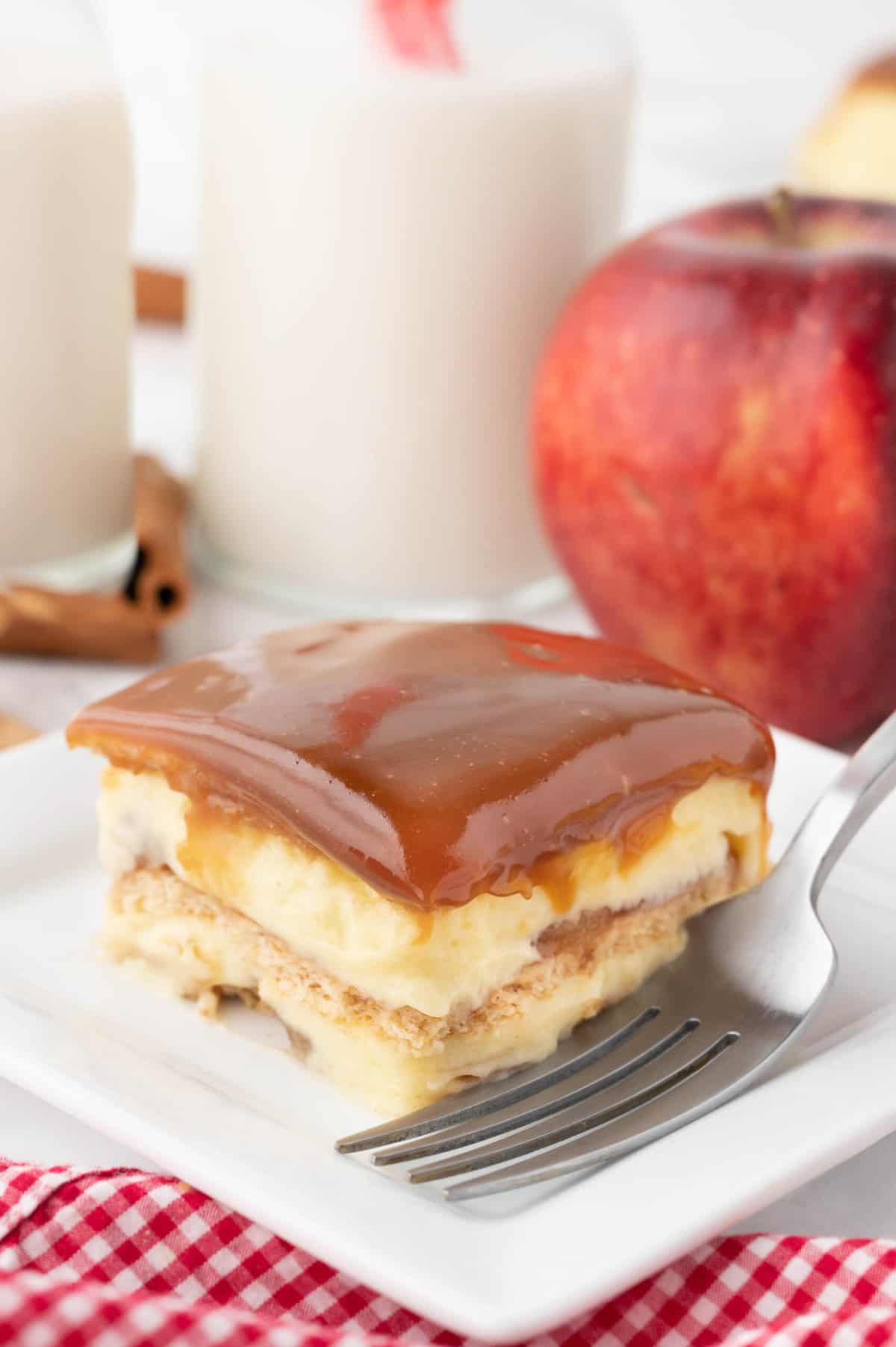 A slice of caramel apple eclair cake on a white plate with a red gingham napkin and a glass of milk next to an apple behind it.