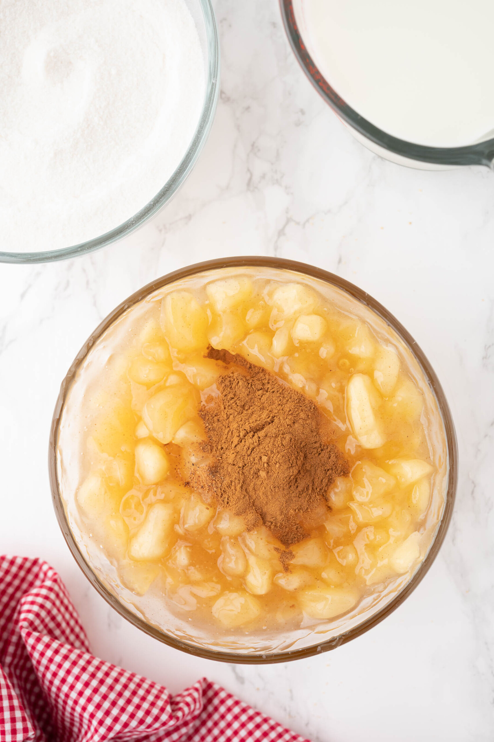 Adding ground cinnamon and nutmeg to apple pie filling in a glass mixing bowl.