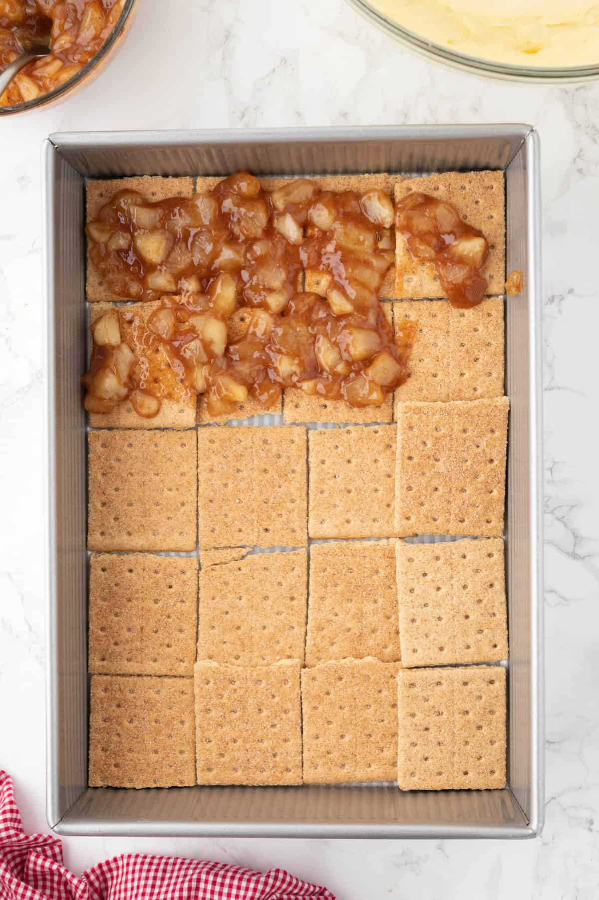 Apple pie filling being spooned over a layer of graham crackers in a rectangular baking dish.