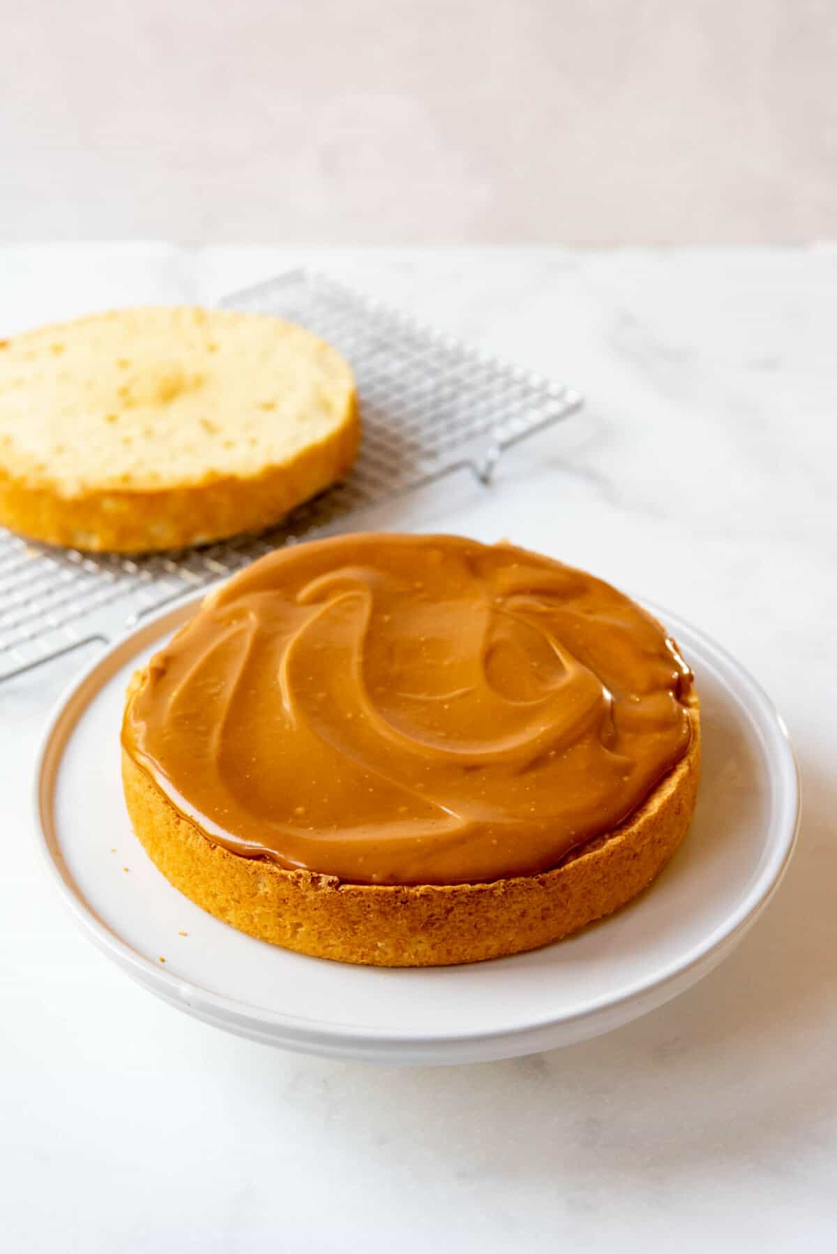 One layer of caramel cake with icing with the second layer on a cooling rack behind it.
