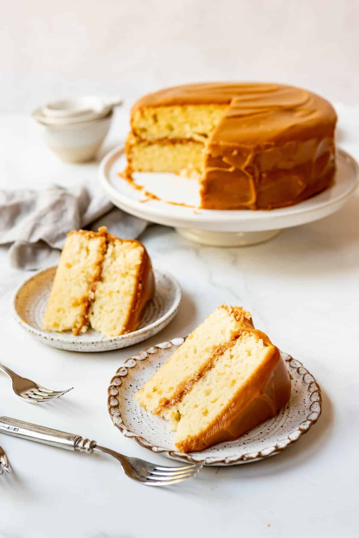 Two slices of caramel cake on plates in front of the rest of the cake on a cake stand.