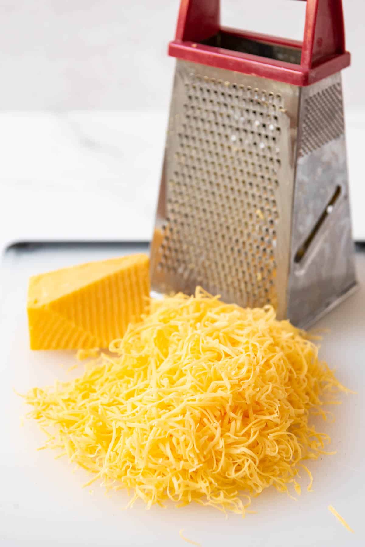 Freshly grated cheddar cheese on a cutting board next to a box grater.
