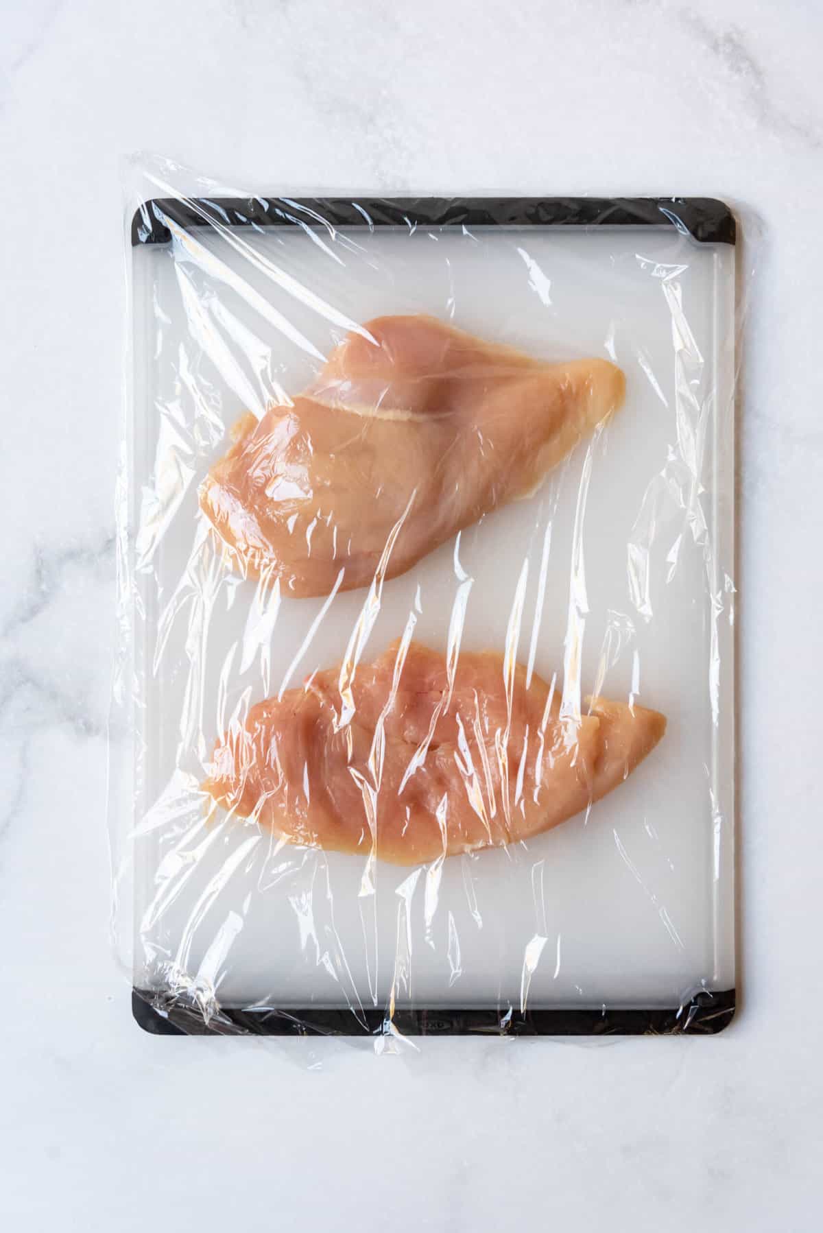 Chicken breasts under a piece of plastic wrap on a cutting board.