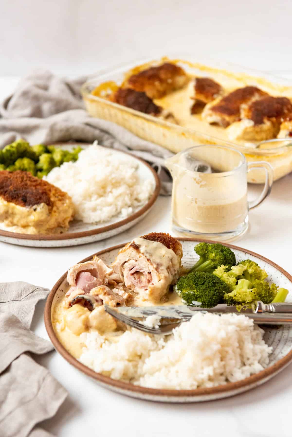 A plate of partially eaten chicken cordon bleu with rice and broccoli next to more plates and a glass pitcher of sauce.