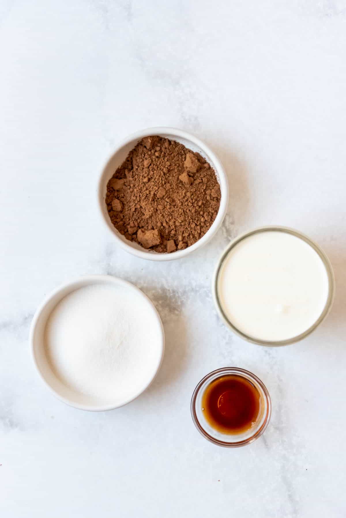 Cocoa powder, sugar, heavy cream, and vanilla extract in separate bowls on a white surface.