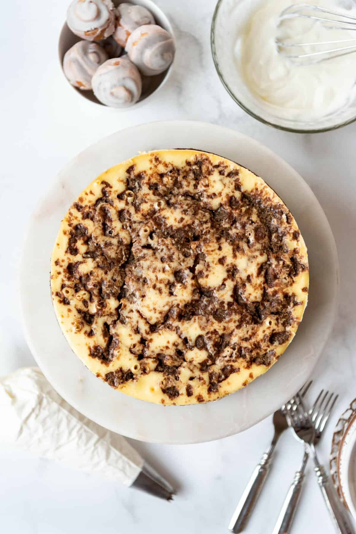 A baked and cooled cinnamon roll cheesecake on a marble cake stand.