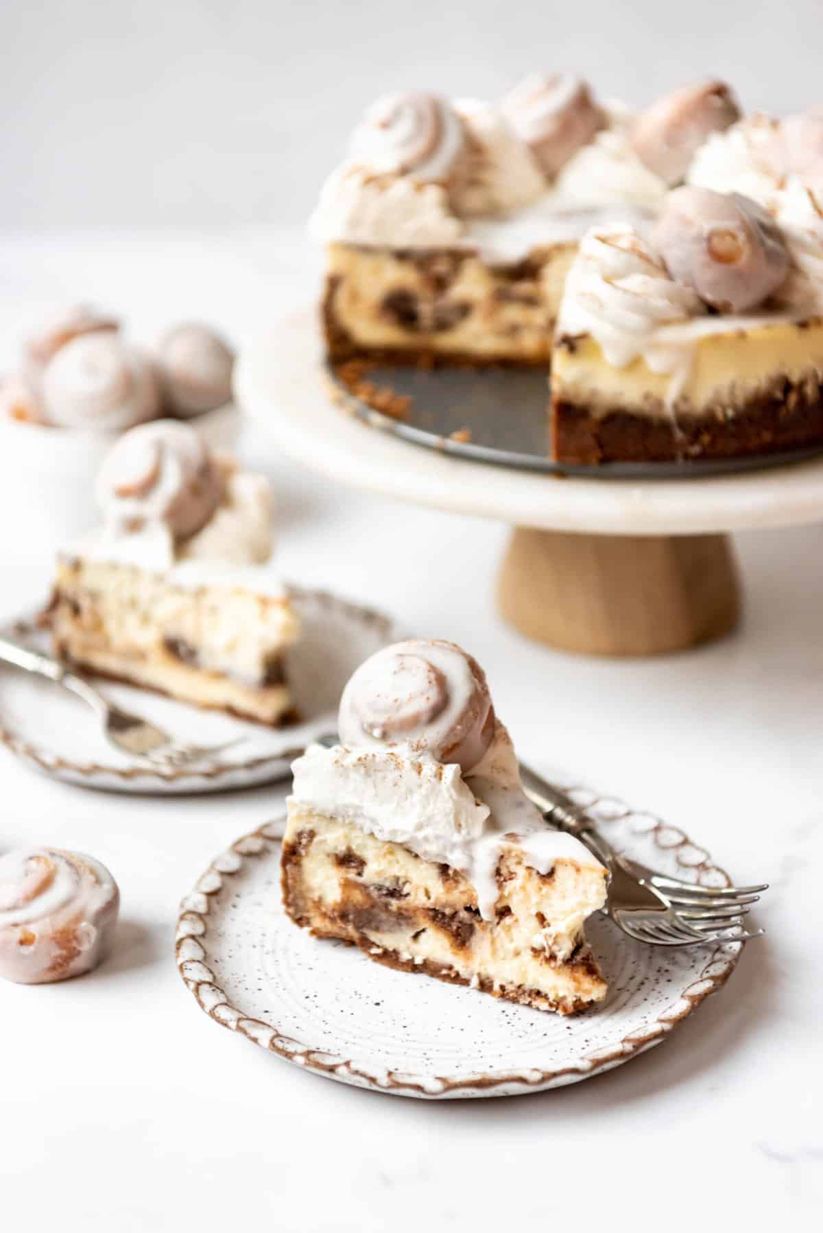 Slices of cinnamon roll cheesecake in front of the rest of the cheesecake on a cake stand in the background.